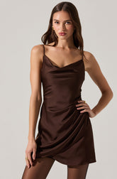 A woman wearing a sleeveless, dark brown satin dress with thin spaghetti straps. The dress features a cowl neckline and a draped, slightly asymmetrical hem.